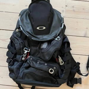 Oakley Tech/Tactical Ryggsäck - Oakley ryggsäck i svart med coola metallspännen, flera fack och detaljer i rött. Väskan har vadderade axelremmar och mesh på baksidan för extra komfort. Passar bra till träning och skolan