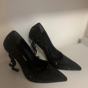 Svarta glittriga YSL pumps med klack - Svarta pumps från Saint Laurent med glittriga stenar över hela skon och en unik klack formad som YSL-loggan. Spetsig tå och hög klack ger en riktigt edgy och lyxig vibe. Perfekta för dig som vill sticka ut med något extra.