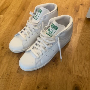 Adidas Stan Smith vita sneakers - Fräscha vita Adidas Stan Smith sneakers i mid cut-modell med gröna detaljer på häl och plös. Klassisk design med snörning, rund tå och perforerade sidor. Skorna är i skinn och har den ikoniska Stan Smith-loggan på baksidan och tungan.        Strl 44