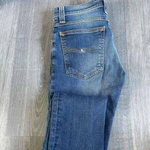Blå  jeans från Nudie Jeans - Snygga blå  jeans från Nudie Jeans med klassisk femficksdesign och ikonisk brodyr på bakfickan. Jeansen har orangea sömmar, dragkedja och en ljus tvätt med slitningar på låren. Perfekta för en avslappnad och trendig look.