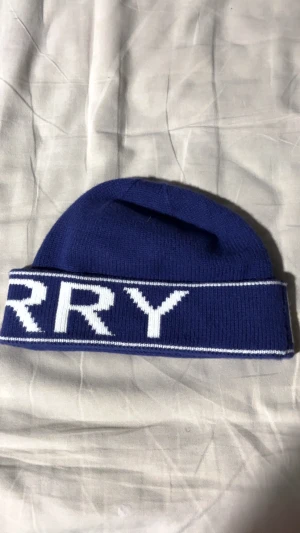 Blå Burberry mössa med vit logga - Snygg marinblå mössa från Burberry med bred uppvikt kant och stor vit BURBERRY-text runt om. Tillverkad i mjuk stickad ull som håller dig varm och ger en clean streetstyle-look. Perfekt för dig som vill ha något exklusivt och trendigt.