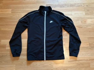 Svart tröja från Nike - Svart tröja med dragkedja / zip från Nike med vit dragkedja och logga på bröstet. Jackan har lång ärm, ståkrage / polo och vita detaljer längs axlarna. Har även byxor till detta set på min sida. 