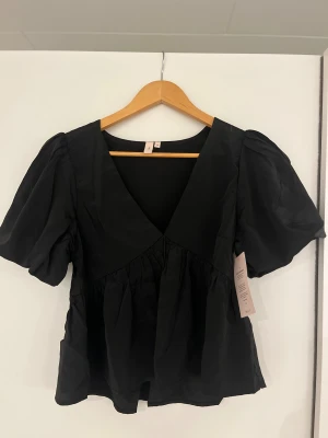 Svart blus från Nelly, strl 36 - Supersnygg svart blus från Nelly med puffiga korta ärmar och djup v-ringning. Modellen har en volangdetalj vid midjan som ger en cool och feminin siluett. Perfekt att styla med jeans eller kjol för en trendig look. Helt oanvänd med lappen kvar.