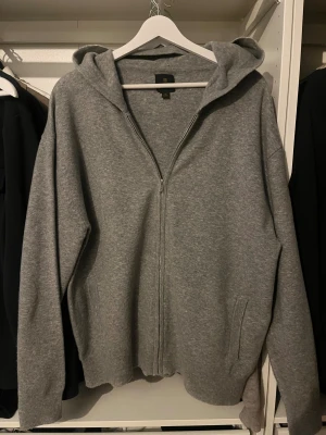 Grå hoodie med dragkedja från NEXT - Grå hoodie från NEXT i storlek M med huva och hel dragkedja framtill. Tröjan har långa ärmar, två fickor och är tillverkad av en mjuk blandning av akryl, nylon, ull och elastan. Perfekt för chill dagar och enkel att matcha med jeans eller joggers.