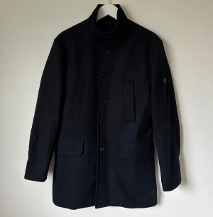 Mörkblå rock från J.Lindeberg, strl 44 - Snygg mörkblå rock från J.Lindeberg i klassisk modell med hög krage och knappar framtill. Jackan har två fickor med lock och två vertikala fickor, samt dekorativ knappdetalj bak. Materialet är 75% ull och 25% polyamid med viskosfoder. Gavin Compact Melton - 4C4