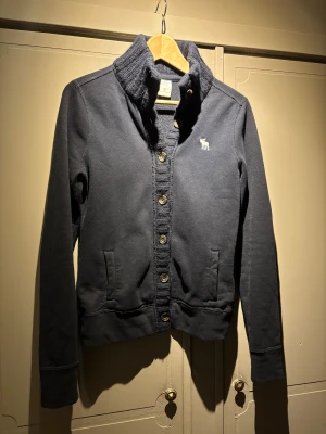 Mörkblå kofta Abercrombie & Fitch - Snygg mörkblå kofta från Abercrombie & Fitch med ribbad krage och knappar hela vägen framtill. Den är liten i storleken så passar även S. Tröjan har ett broderat vitt älg-logo på bröstet, två fickor och är tillverkad i mjuk bomull med stickade detaljer vid kragen och knappslån. Perfekt för chill höstkvällar.