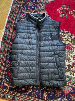 Svart quiltad dunväst från BOSS - Svart dunväst från BOSS med quiltad design och hög krage. Västen har dragkedja framtill, två snedställda fickor med knappstängning och en bred BOSS-logga på bröstet. Materialet är lätt och syntetiskt, perfekt för lager-på-lager. Snygg detalj med kontrastband i kragen.