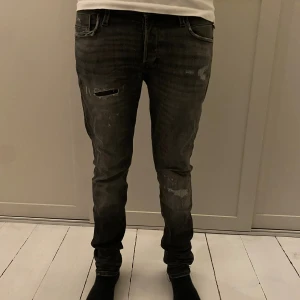 Jack and Jones svarta/gråa jeans med slitningar - Säljer ett par grå/svarta Jack and Jones med coola slitningar och detaljer på både fram- och baksida. Jeansen är slim fit och både väldigt sköna och feta. Storleken är 29/32