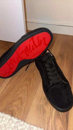 Christian Louboutin svarta sneakers - Svarta sneakers från Christian Louboutin med ikonisk röd sula och snygg signatur. Skorna har rund tå, platt sula och snörning. Ovandelen är i mjuk mocka och insidan är beige med guldfärgad logga. Perfekt för dig som gillar lyxiga detaljer och stilren design.