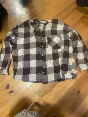 Rutig henley-skjorta från H&M Divided - Säljer en chill rutig henley-skjorta från H&M Divided i storlek S. Skjortan har bruna, gråa och vita rutor, lång ärm och en bröstficka. Knappar framtill och mjukt bomullsmaterial gör den skön att bära. Perfekt för dig som gillar avslappnad stil.