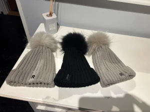 Rockandblue och Hollies ribbstickad mössa med pälsboll  - Säljer snygga ribbstickade mössor från Rockandblue med stor fluffig pälsboll på toppen. Finns i svart med svart boll och beige med ljusgrå boll. Perfekt accessoar för kalla dagar, både varm och trendig med diskret logga framtill.