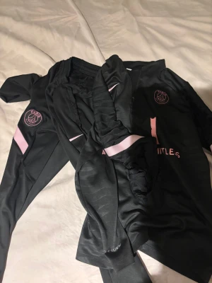 Svart PSG fotbollströja med rosa detaljer - Säljer en svart Paris Saint-Germain fotbollströja från Nike med rosa logga och detaljer. Tröjan har lång ärm och är tillverkad i ett lätt, sportigt material som andas. Perfekt för dig som vill ha en snygg och modern look på planen.
