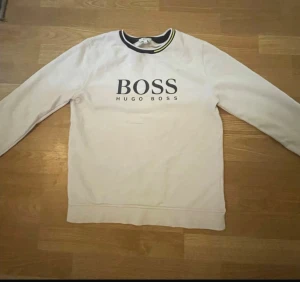 Vit sweatshirt från Hugo Boss - Vit sweatshirt från Hugo Boss med svart BOSS-tryck på bröstet. Rund hals med svart och gul detalj, samt ribbade muddar vid hals, ärmslut och nederkant. Långärmad och i mjukt bomullsmaterial, perfekt för en clean och stilren look.