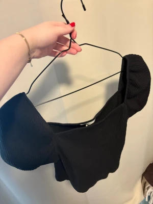 Svart croppad offshoulder topp - Säljer en svart croppad topp med offshoulder-design och puffiga ärmar. Toppen är ribbad och har en snygg, figurnära passform som ger en cool vibe. Perfekt att styla med höga jeans eller kjol för en trendig look.
