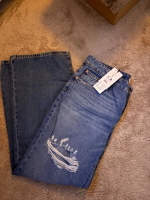 Blå jeans med slitning och tagg - Snygga blå jeans med en cool sliten detalj på ena benet och klassisk femficksdesign. Jeansen har raka ben och en avslappnad passform, perfekta för en trendig streetstyle-look. Materialet är robust denim och färgen är mellanblå.