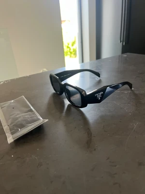 Svarta solglasögon från Prada - Snygga svarta solglasögon från Prada med breda skalmar och tydlig logga på sidan. Glasögonen har en modern, kantig form och är tillverkade i blankt syntetmaterial. Perfekta för dig som vill ha en stilren och exklusiv look. OBS endast glasögonen medföljs. Putsduk borttappad 