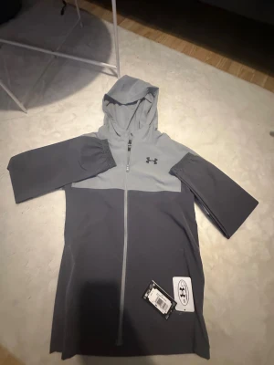 Under Armour Jacka - Snygg tvåtonad grå vindjacka från Under Armour med huva och hel dragkedja framtill. Jackan har långärmade ärmar och logga på bröstet. Perfekt för träning eller chill dagar ute. Modern och sportig look med stilren design.