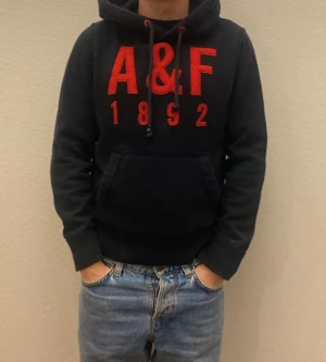 Svart hoodie Abercrombie & Fitch M - Svart hoodie från Abercrombie & Fitch med stor röd A&F-logga och siffror 1892 på bröstet. Tröjan har huva med snörning, magficka och långa ärmar. Mjuk insida och klassisk streetstil, perfekt för chill dagar.
