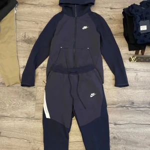 Nike tech fleece old season - mörkblå tracksuit från Nike med vit logga på benet. Byxorna har dragsko i midjan och en sportig look med paneler i olika blå nyanser och vita detaljer på sidan. Perfekta för chill eller träning. Nike Tech Old Season. Ett litet hål som man kan sy in, har inte använts