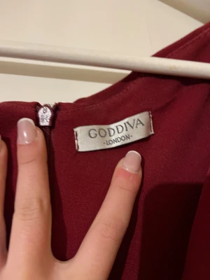 Vinröd långklänning från Goddiva - Säljer en elegant vinröd långklänning från Goddiva London med omlott-detalj framtill och v-ringad hals. Klänningen är ärmlös och har en figurnära passform som framhäver siluetten. Materialet är mjukt och har en lätt stretch. Perfekt för bal eller fest! Klänningen är upp sydd lite men inget som syns 