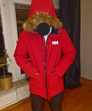 Röd vinterjacka från Helly Hansen med fuskpäls - Riktigt snygg röd vinterjacka från Helly Hansen med stor huva och fuskpäls. Jackan är vadderad, har dragkedja och knappar framtill samt två stora fickor. Perfekt för kalla dagar och har en clean, sportig vibe. Knappt använd och nypriset är 2500kr