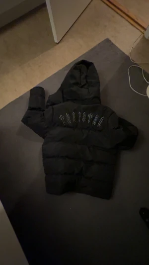 Svart pufferjacka från Trapstar L - Svart pufferjacka från Trapstar med broderad logga på ryggen och huva. Jackan har dragkedja framtill och coola detaljer som metallbadge på armen.