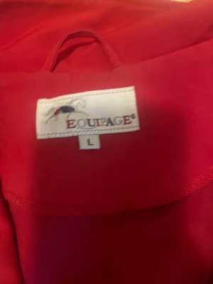 Röd vindjacka från Equipage, stl L - Snygg röd vindjacka från Equipage med broderad logga på bröstet och ett coolt märke på ärmen. Jackan har två fickor med dragkedja och en stilren ståkrage. Perfekt för dig som gillar sportig och enkel stil.