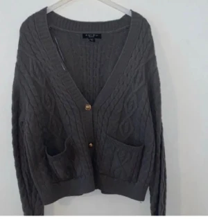 Grå stickad kofta med fickor - Mysig grå stickad kofta från AWARE med snyggt kabelmönster, två framfickor och guldfärgade knappar. Koftan har v-ringad hals och långa ärmar, perfekt för lager-på-lager. Passar dig som gillar chill och stilren look.