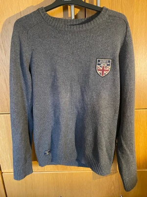 Grå stickad tröja Morris Gents M - Snygg grå stickad tröja från Morris Gents i storlek medium. Tröjan har rund hals, ribbade muddar och två coola patches – en med Union Jack och en med Morris Gents-logga. Perfekt för dig som gillar klassisk stil med brittisk touch.