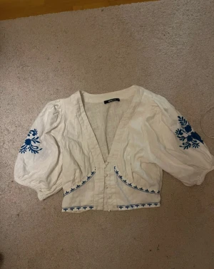 Vit blus med blå broderi och puffärm - Säljer en vit blus från Gina med blå broderade detaljer på ärmar och längs kanten. Blusen har puffiga trekvartsärmar, v-ringning och knappar framtill. Perfekt för dig som gillar boho-vibe och snygga detaljer.