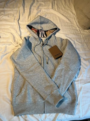 Grå hoodie från Burberry med dragkedja - Snygg grå hoodie från Burberry med klassiskt rutigt foder i huvan och broderad logga på bröstet. Tröjan har snörning i huvan, ribbade muddar och dragkedja framtill. Perfekt för dig som vill ha en stilren och lyxig look.