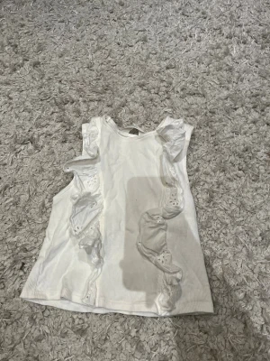Vit ribbad linnetopp med volanger H&M - Säljer en vit ribbad linnetopp från H&M i storlek 146/152. Toppen är ärmlös och har snygga volangdetaljer med broderade inslag längs axlarna och sidorna. Perfekt för dig som gillar en söt och trendig look med extra detaljer.