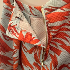 Vacker mönstrad kjol från H&M - Midilång kjol från H&M med ett tropiskt orange mönster på ljusblå bas. Kjolen har en A-linjeformad siluett, markerad midja och guldfärgad dragkedja bak. Materialet har en lyxig lite styv struktur som håller formen,  och modellen med fickor ger en cool känsla. Färgkombinationen är perfekt och passar super till vitt eller svart. Funkar året om beroende på hur den stylas. Modellen på bilden är strl 36/S och kjolen passar bra både till S/M. 