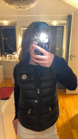 Svart väst från Moncler - Svart dunväst från Moncler. Västen har två fickor med dragkedja och klassisk Moncler-logga på bröstet. Väldigt bra skick då den har varit väl bähandlad. Storlek 12/152cm. Du får även en orginal påse till västen som man kan rulla ihop och stoppa in.