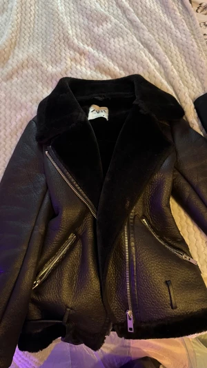 Svart pälsjacka med bälte från Zara - Säljer en svart pälsjacka från Zara med bred krage, synliga silvriga dragkedjor och bälte i midjan. Jackan har mjukt pälsfoder och två fickor med dragkedja. Perfekt för dig som vill ha en cool och varm look under höst och vinter.