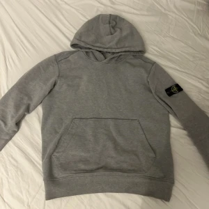 Grå Stone Island hoodie med huva - Snygg grå hoodie från Stone Island med klassisk patch på vänster ärm. Tröjan har huva, magficka och ribbade muddar. Perfekt för dig som gillar streetwear och vill ha en stilren look. Materialet är mjukt och skönt, troligen bomull eller bomullsblandning. Pris kan diskuteras!