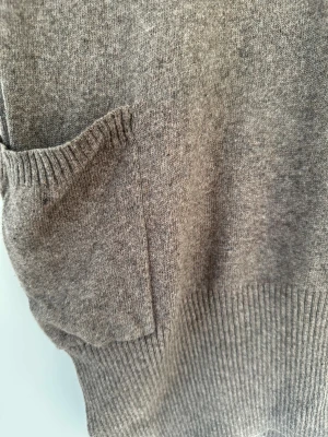 Brun stickad polotröja / klänning - Mysig brun stickad polotröja med korta ärmar och två praktiska fickor framtill. Tröjan har en bred, ribbad polokrage och knappar längs ryggen för extra detalj. Perfekt för lager-på-lager under höst och vinter.