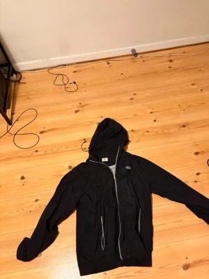 Svart vindjacka från Lacoste Sport - Svart vindjacka från Lacoste Sport med huva och dragkedja framtill. Jackan har två fickor med dragkedja och den klassiska gröna krokodilloggan på bröstet. Lätt och smidig design i syntetmaterial, perfekt för blåsiga dagar.