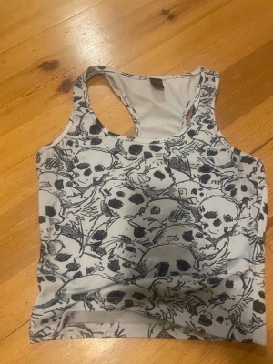 Vit linnetopp med döskallemönster Romwe XS - Cool vit linnetopp från Romwe i storlek XS, täckt med svart döskallemönster. Modellen är ärmlös med brottarrygg och har en slim passform. Perfekt för dig som gillar edgy och unik stil.