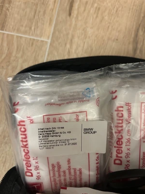 Svart första hjälpen-väska BMW - Praktisk svart första hjälpen-väska från BMW Group med vit text och kors-symbol på framsidan. Väskan är rektangulär med dragkedja och innehåller förbandsmaterial enligt DIN 13164. Perfekt att ha i bilen för extra trygghet.