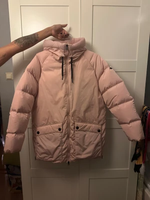 Ljusrosa Peak Performance stella jacket  - Säljer en ljusrosa pufferjacka från Peak Performance i storlek S. Jackan har luva, dragkedja framtill, två stora fickor med knappar. Perfekt för kalla dagar med sin oversized vibe och snygga look.