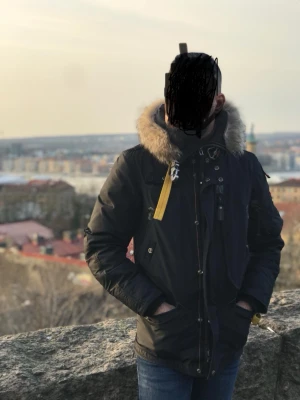 Svart Parajumpers parkas med päls - Säljer en svart Parajumpers parkas med ikonisk gul rem, flera fickor och detaljerade dragkedjor. Jackan har en stor huva med äkta päls och patch på ärmen. Perfekt för kalla dagar. Det gar även att ta av fodret pa insidan för att kunna använda jackan pả höst/var. Jackan är i bra skick