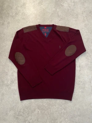 Vinröd stickad tröja Polo Ralph Lauren - Säljer en vinröd stickad tröja från Polo Ralph Lauren med bruna quiltade detaljer på axlar och armbågar. Klassisk v-ringning och broderad logga på bröstet. Perfekt för höst och vinter, riktigt snygg till jeans eller chinos.