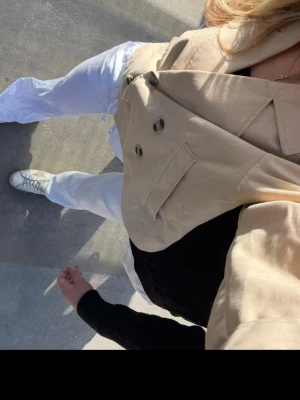 Beige croppad trenchcoat  - Säljer en beige croppad trenchcoat med dubbelknäppning och stora slag. Jackan har axelklaffar, knappar vid ärmslut och två fickor framtill. Perfekt för dig som vill ha en trendig och stilren look till våren eller hösten.