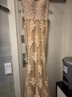 Champagnefärgad långklänning med spets - Säljer en elegant långklänning i champagnefärg med detaljerad spets och pärlor upptill. Klänningen har figurnära passform, är ärmlös och dekorerad med blommiga spetsapplikationer över hela tyget. Perfekt för bal eller festliga tillfällen.