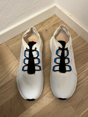 Vita sneakers från Hummel - Säljer ett par vita sneakers från Hummel med svarta och blå detaljer. Skorna har svart snörning, vit ovandel i syntetmaterial och en chunky sula med svart och blått mönster undertill. Sidan pryds av Hummels klassiska pilar i vitt och röd text.