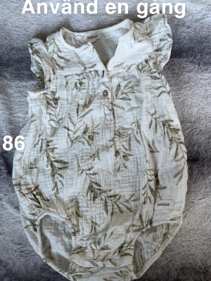 Blommig body från Newbie - Supersöt body från Newbie i vitt med grönt blommönster. volangdetaljer vid axlarna och knappar framtill. Perfekt för vår och sommar, lätt och luftig för små barn.🌸 Använd en gång. Storlek 86