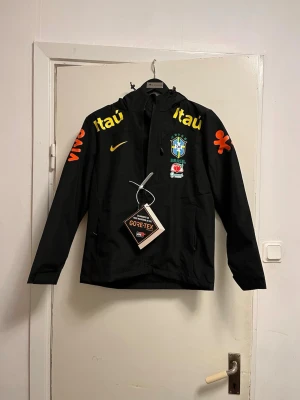 Svart Brazil Nike vindjacka Gore-Tex - Svart vindjacka från Nike med Brazil-logga och gula samt orangea tryck. Jackan har huva, dragkedja och är tillverkad i Gore-Tex-material. Perfekt för dig som gillar sportig stil och vill visa stöd för Brasilien. Snygga detaljer med sponsorer på bröst och ärmar. Finns i storlek S-M-L 