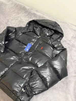 Ralph Lauren jacka  - Svart, glansig pufferjacka från Polo Ralph Lauren med huva och dragkedja framtill. Jackan har två sidofickor med dragkedja och en röd broderad logga på bröstet. Perfekt för kalla dagar och har en riktigt snygg, modern look.