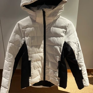Vit och svart pufferjacka från Everest - Snygg pufferjacka från Everest i vit och svart med huva och dragkedja framtill. Jackan har två fickor med dragkedja och är quiltad för extra värme. Perfekt för kalla dagar och har en sportig look med kontrastpaneler på sidor och ärmar.